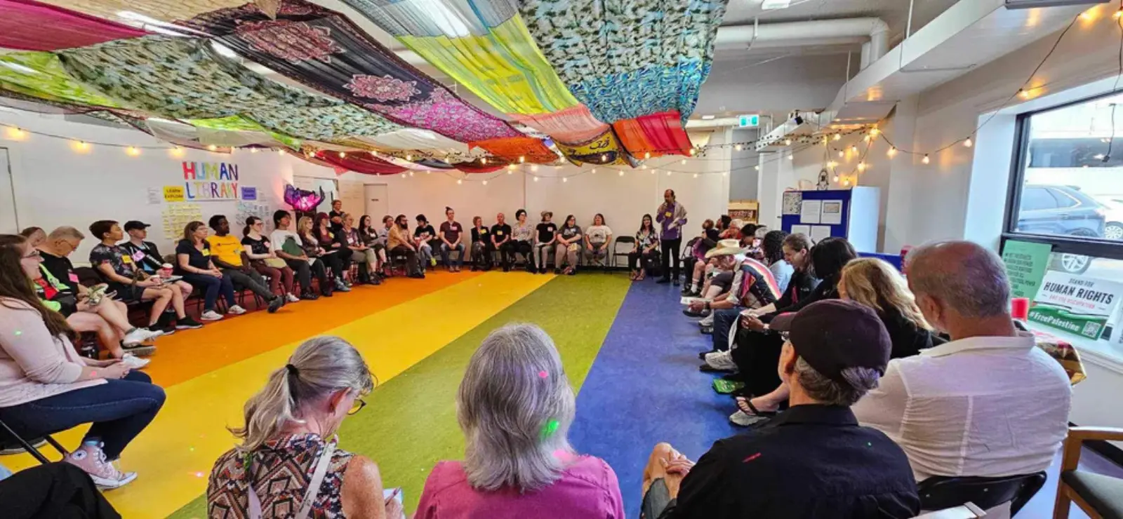 People sitting in a wide circle on the rainbow floor at 206 Piccadilly, under a ceiling draped with fabric